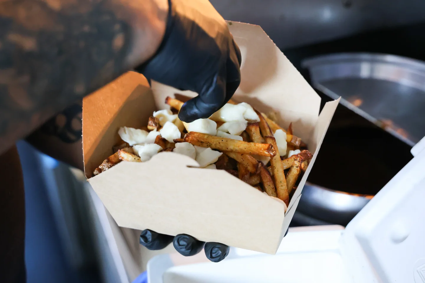 Tattooed hand adding cheese curds to fresh-cut fries in a kraft box
